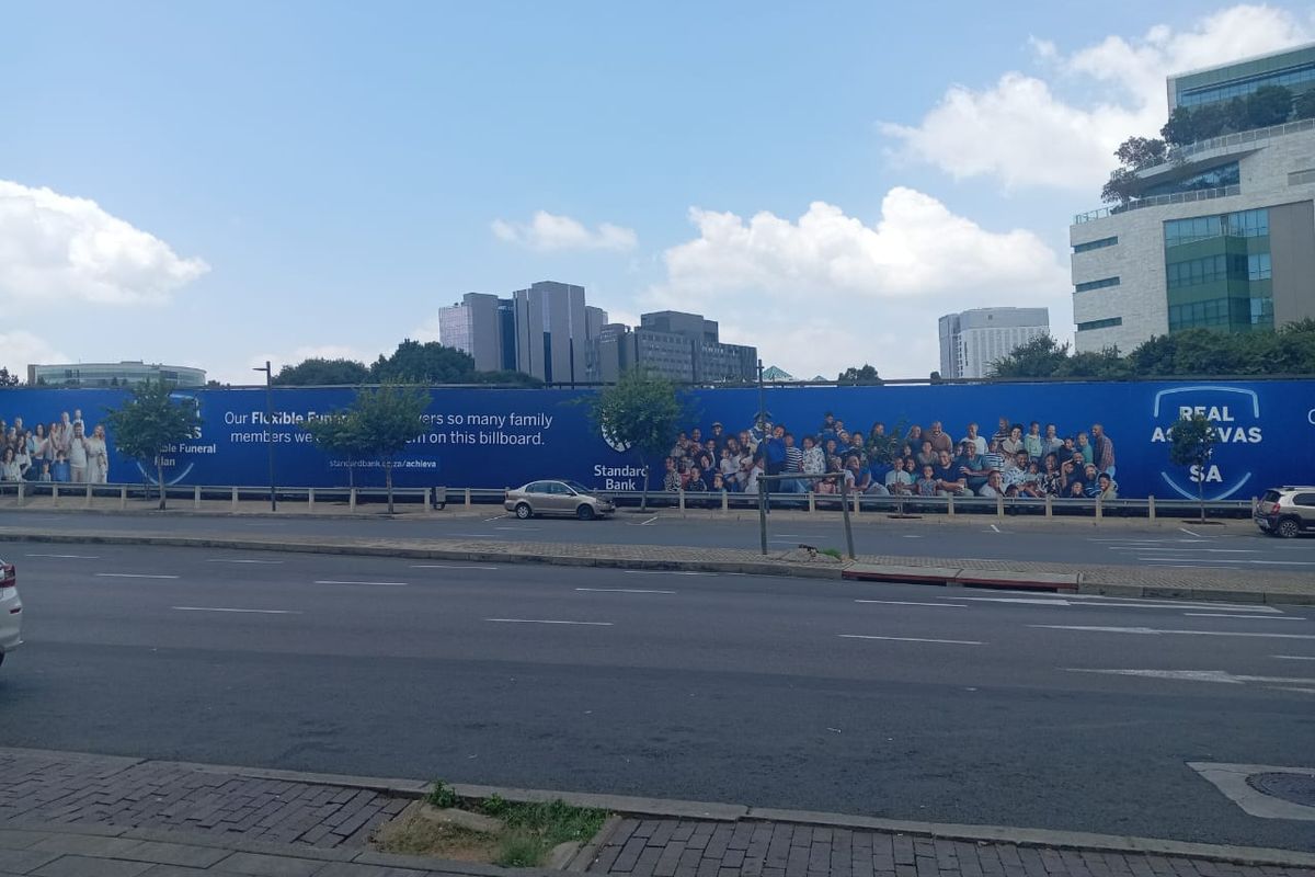A street-level view showing a large advertising billboard situated along a public road, with urban buildings visible in the background.