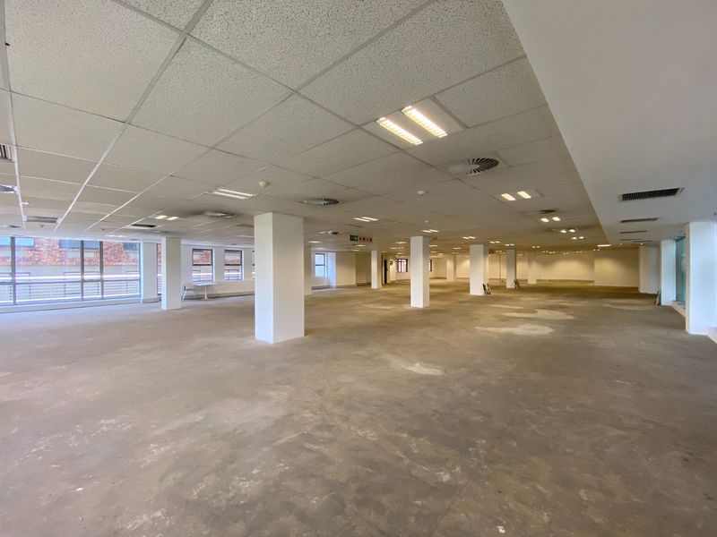 A large, open-plan commercial office space featuring structural columns, suspended ceilings with recessed lighting, and expansive windows providing natural light.
