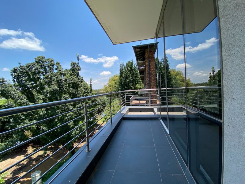 A modern, spacious balcony featuring sleek stainless steel railings, dark tiled flooring, and expansive glass windows overlooking a lush green landscape.