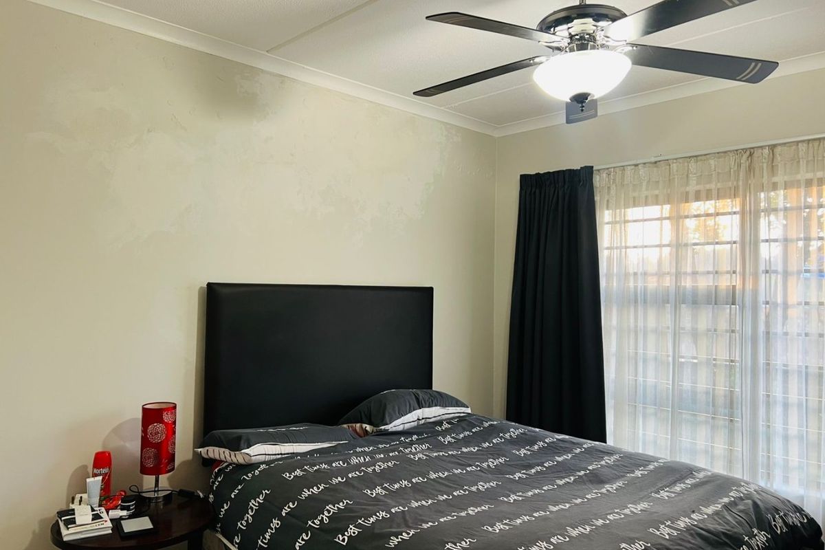 A bedroom featuring a ceiling fan and window security bars, with walls showing signs of uneven paint application.