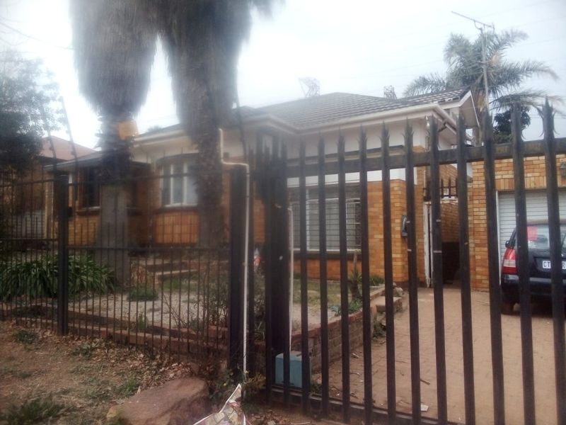 Front view of a single-story brick house featuring a tiled roof, paved driveway, and secure metal perimeter fencing.