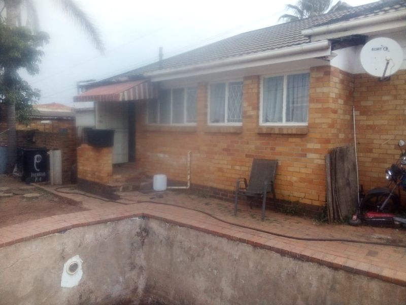 Rear exterior view of a brick house featuring a swimming pool with visible wear and a small covered entrance area.