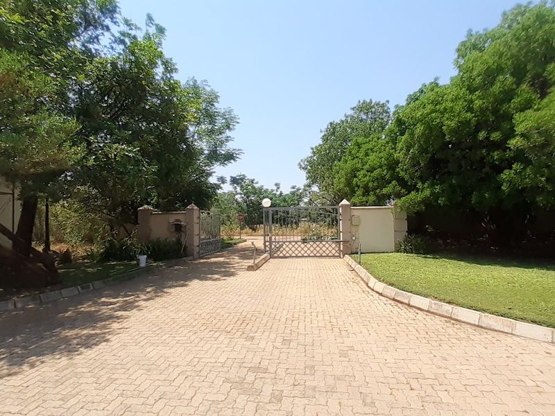 A well-maintained residential entrance featuring a paved driveway, metal security gate, and established landscaping with mature trees.