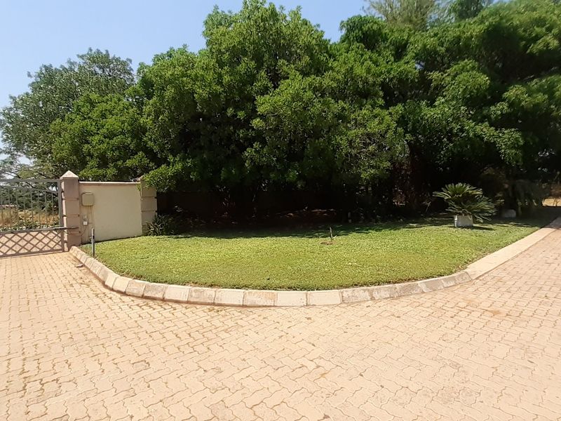 A view of the property entrance featuring a paved driveway, a metal security gate, and a landscaped garden area with mature trees.