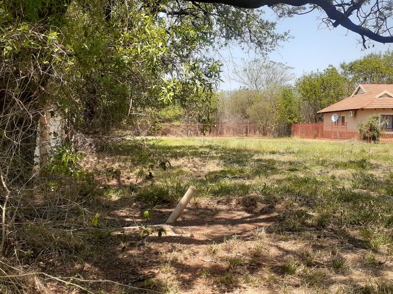 A view of the residential vacant land featuring mature trees, natural grass coverage, and perimeter fencing with an adjacent dwelling visible.