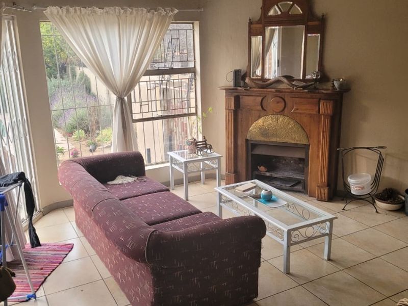 A living room featuring a traditional wooden fireplace, tiled flooring, and exposed beam ceilings with natural light from large windows.