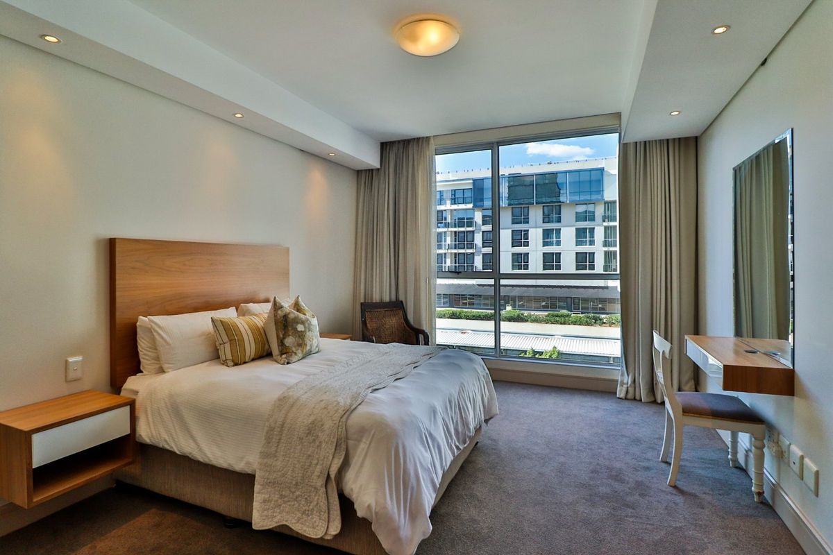 A well-maintained bedroom featuring neutral tones, carpeted flooring, recessed lighting, and a large window offering an urban view.