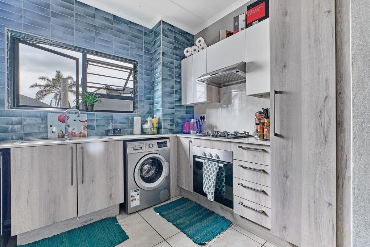 A compact, modern kitchen featuring light wood-grain cabinetry, blue tiled splashbacks, a gas hob, built-in oven, and integrated laundry appliance space.