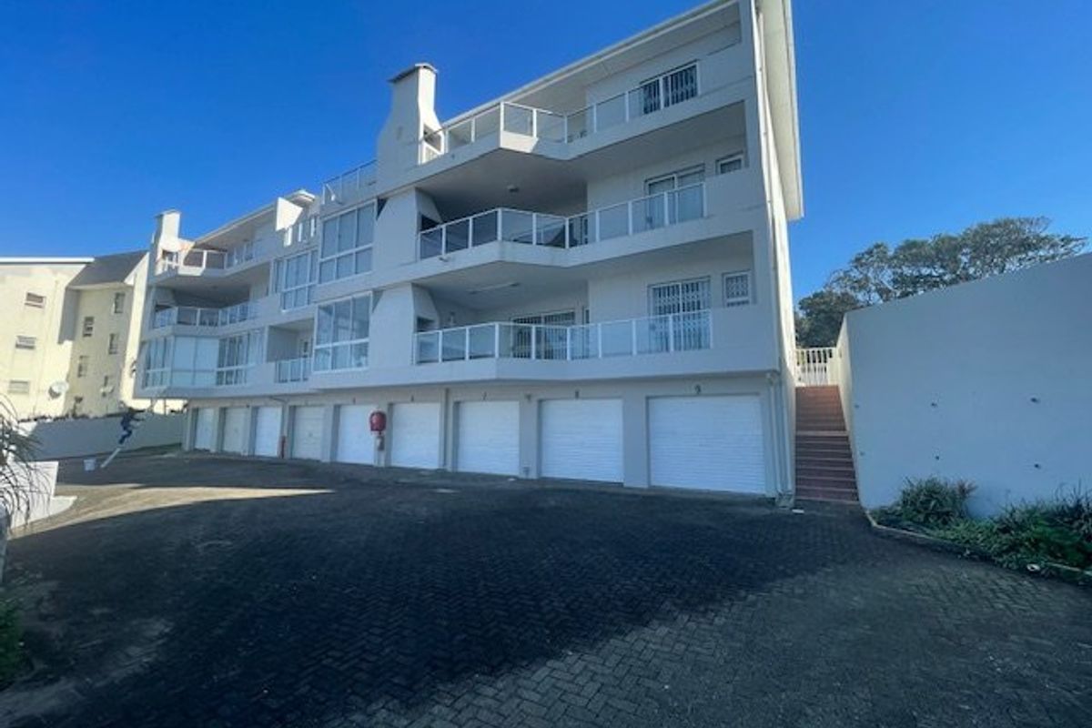 Rear view of a multi-story apartment complex featuring multiple ground-level garages, private balconies with glass balustrades, and a paved driveway area.