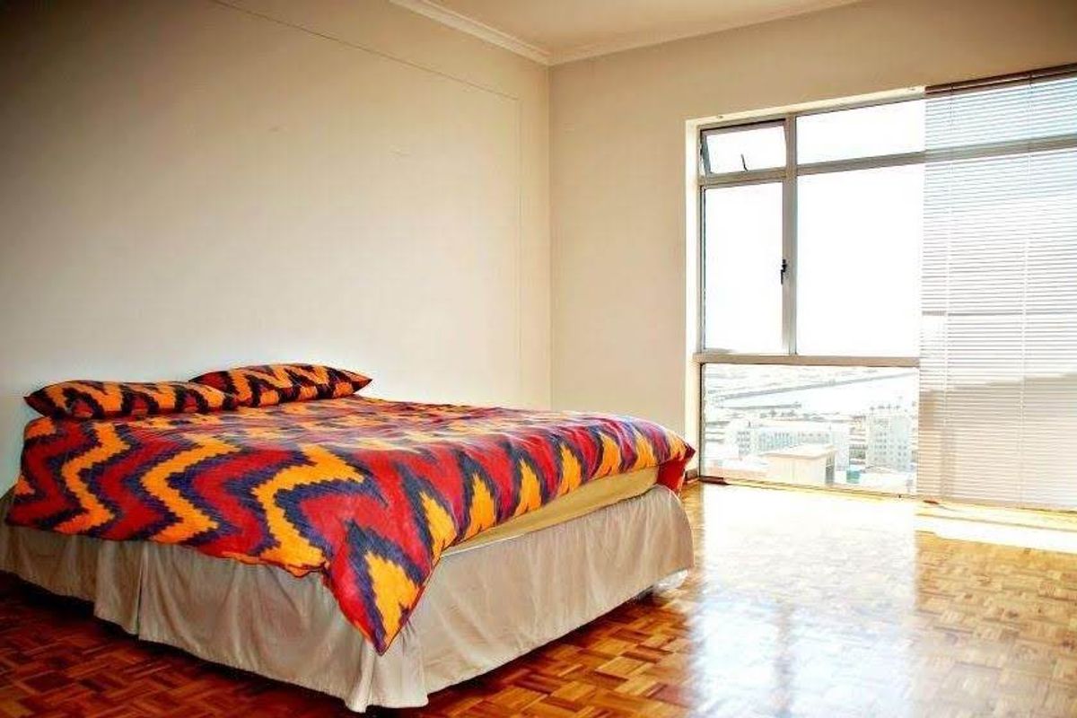 A bedroom featuring classic parquet flooring and a large window with venetian blinds, offering natural light and an urban view.