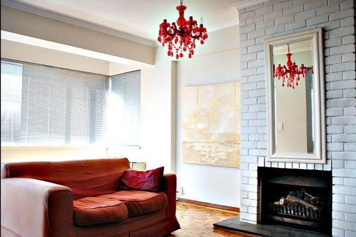 A cozy living room featuring a painted brick fireplace, parquet flooring, and large windows with venetian blinds, offering a bright and inviting atmosphere.