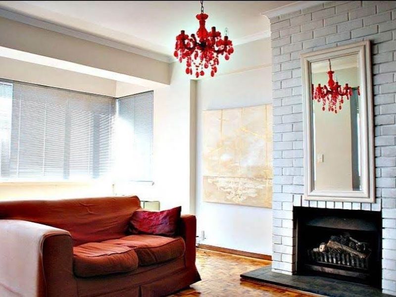A cozy living room featuring a painted brick fireplace, parquet flooring, and large windows with venetian blinds, offering a bright and inviting atmosphere.