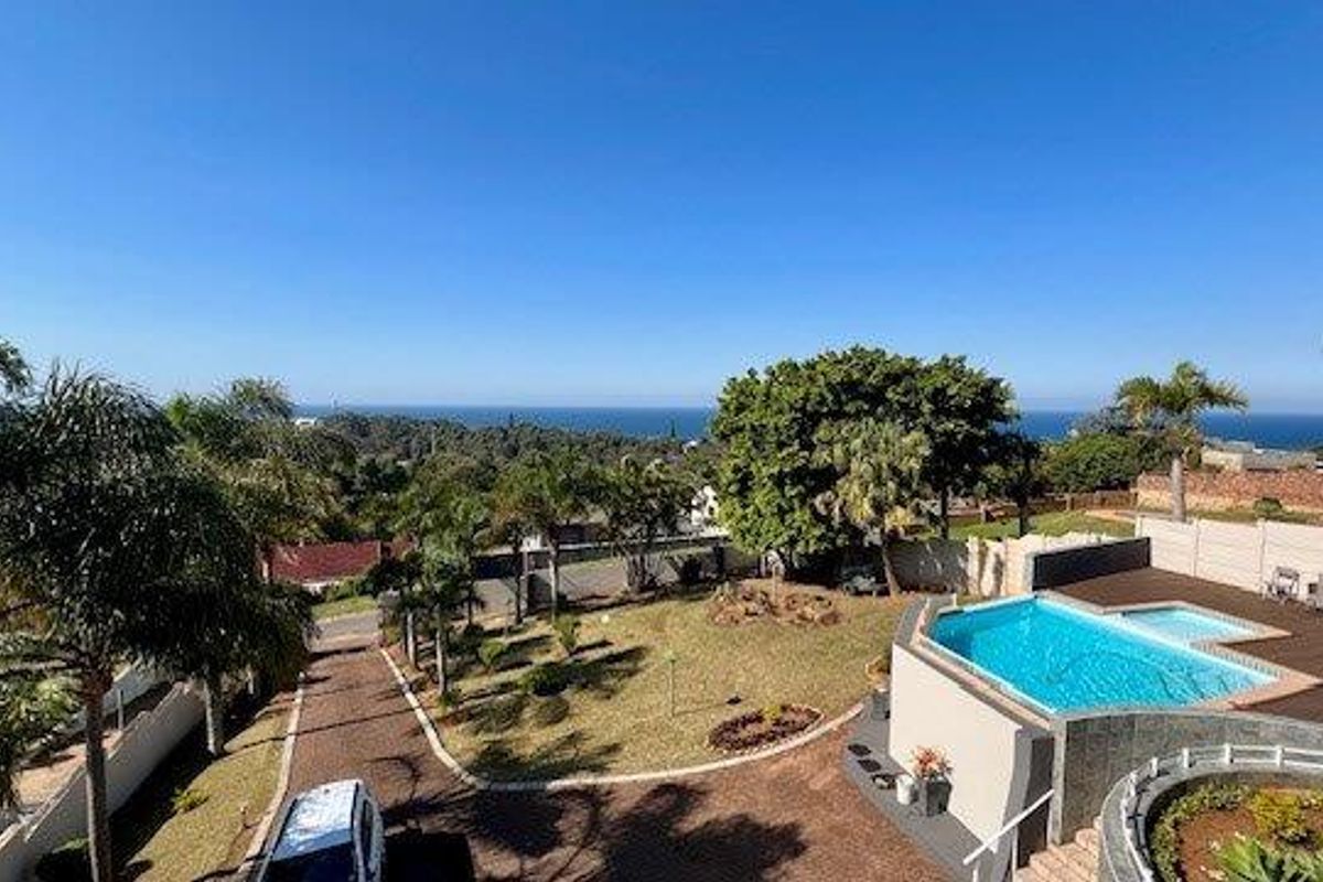 Aerial view of the property featuring a swimming pool, landscaped garden, paved driveway, and distant ocean views.