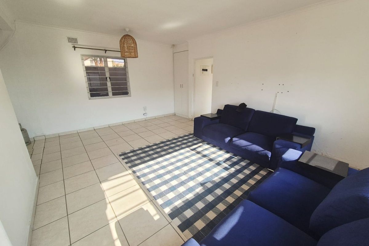 A spacious living room featuring tiled flooring, a built-in cupboard, and natural light through a window with security bars.