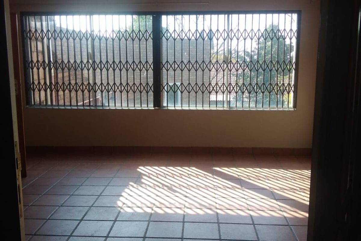 An empty living room featuring terracotta-style floor tiles, a glossy ceiling finish, and a large window secured with a metal security gate.