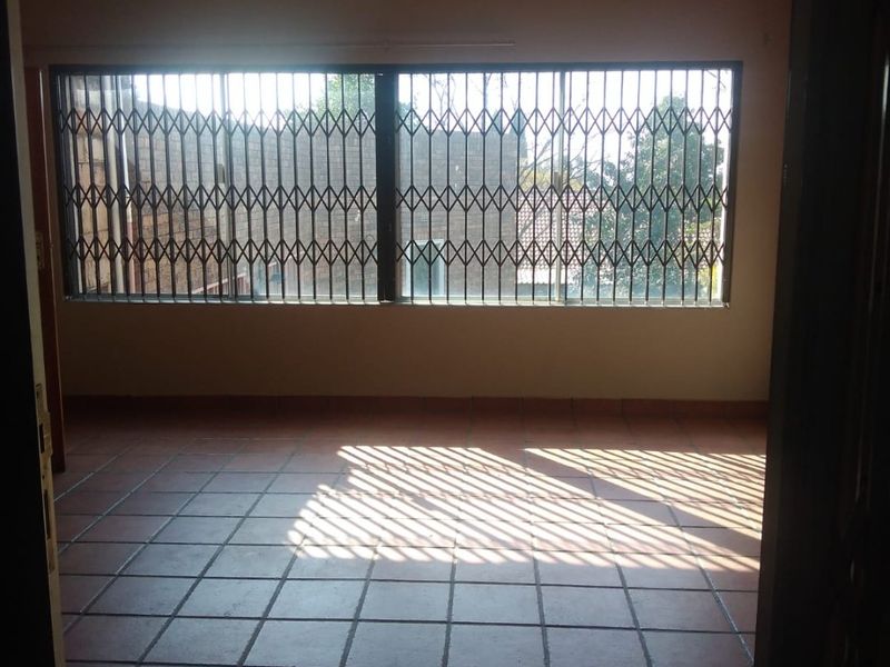 An empty living room featuring terracotta-style floor tiles, a glossy ceiling finish, and a large window secured with a metal security gate.