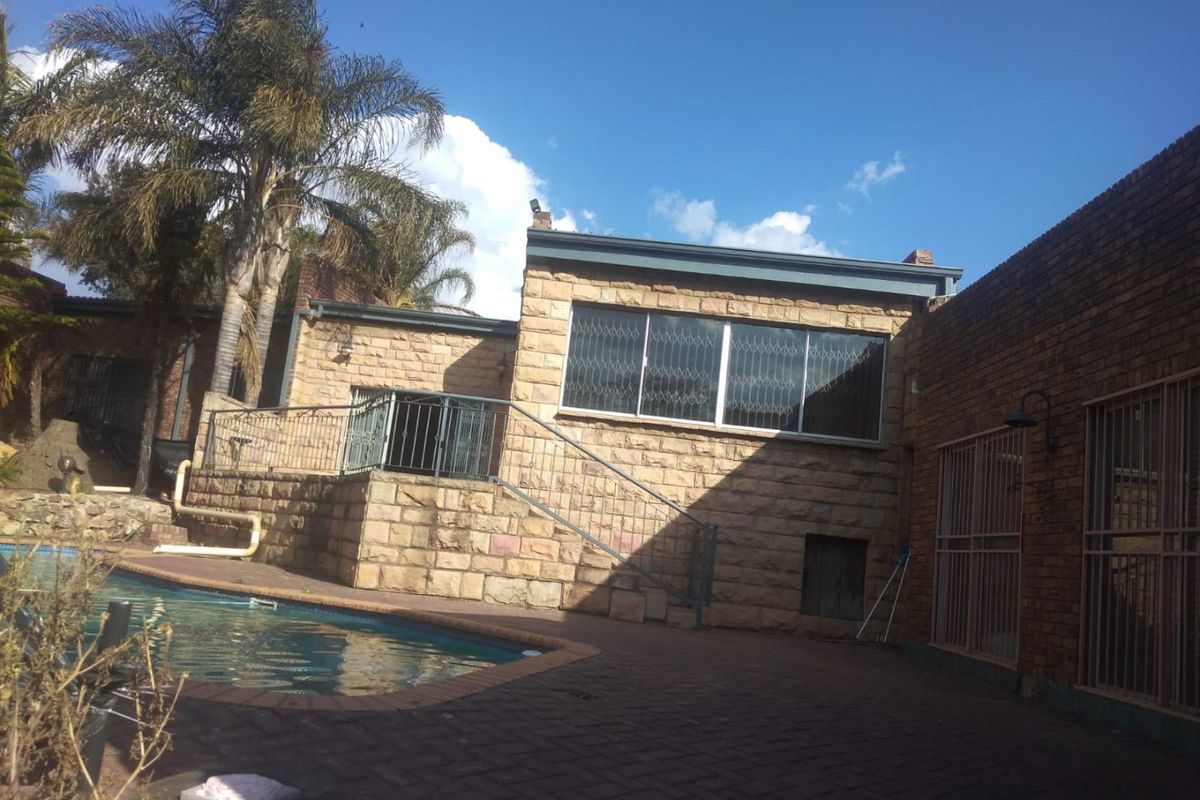 Rear exterior view of the property featuring a swimming pool, stone-clad facade, and paved patio area with security features.