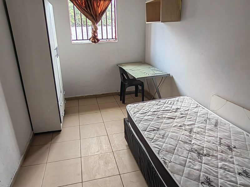 A compact bedroom featuring tiled flooring, a window with security bars, and wall-mounted shelving, offering a functional space in need of cosmetic updates.