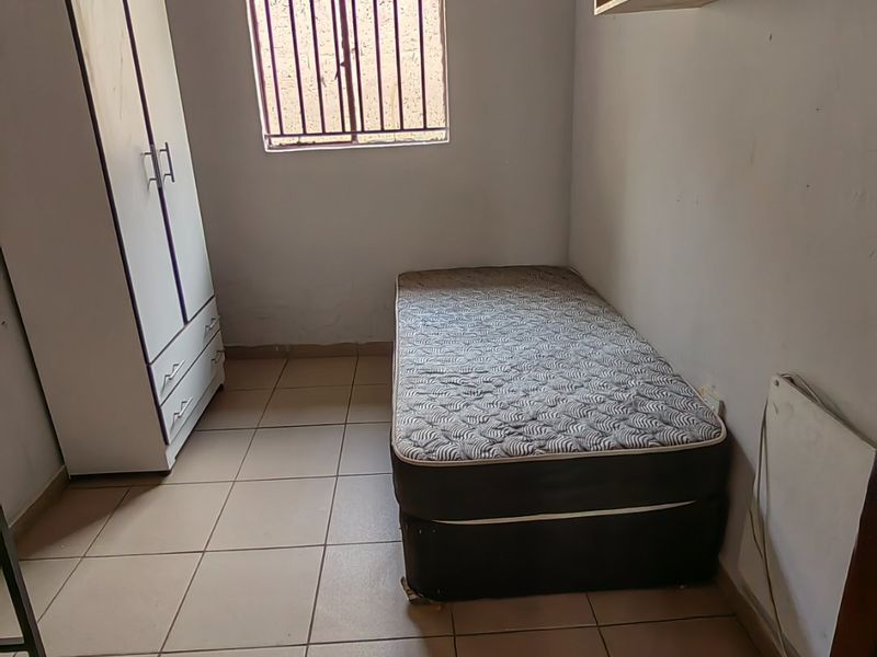 A compact bedroom featuring tiled flooring, security bars on the window, and built-in shelving, requiring cosmetic updates to the walls and ceiling.