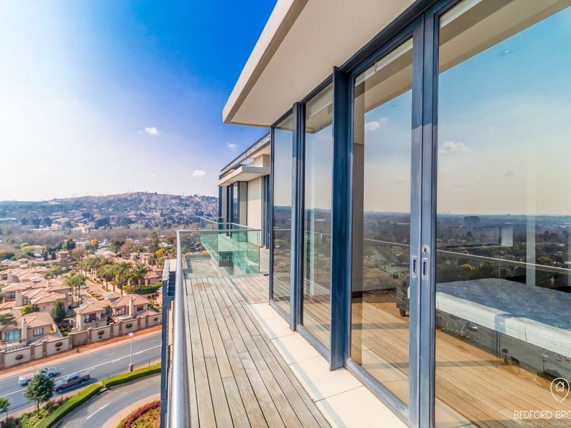 A modern, elevated balcony featuring timber decking, a sleek glass balustrade, and expansive sliding glass doors offering panoramic views of the surrounding area.