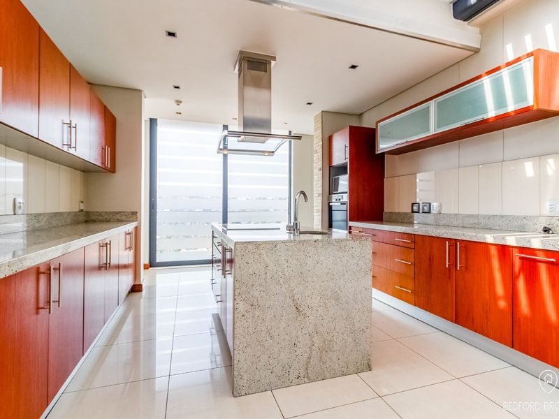Modern kitchen featuring warm wood cabinetry, granite countertops, a central island with sink, and built-in appliances, illuminated by natural light from large windows.