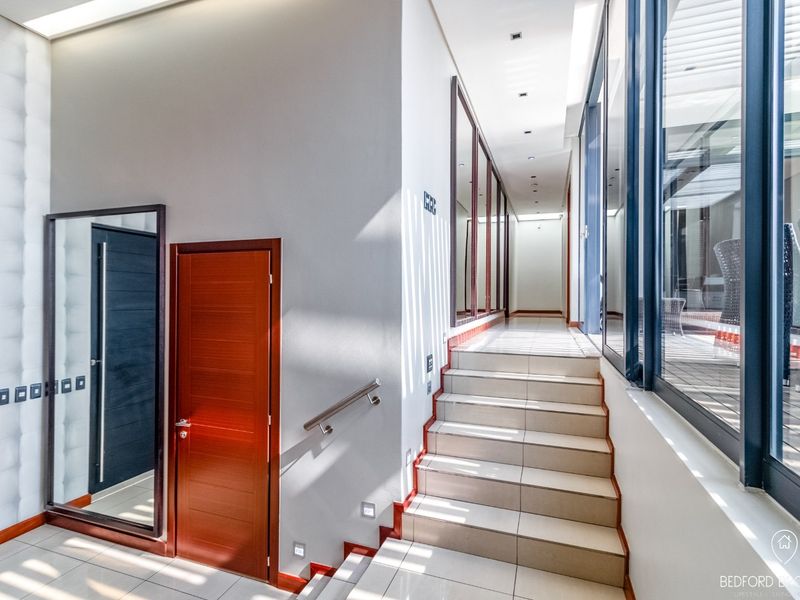 A bright, modern hallway featuring tiled flooring, a staircase with a stainless steel handrail, and large glass windows overlooking an outdoor area.