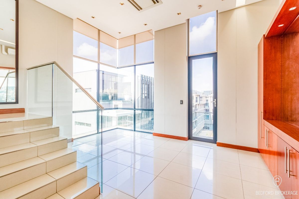 A bright, modern living area featuring large floor-to-ceiling windows, tiled flooring, a glass balustrade staircase, and sleek built-in cabinetry.