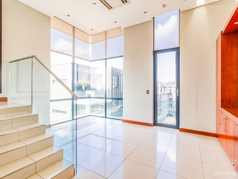 A bright, modern living area featuring large floor-to-ceiling windows, tiled flooring, a glass balustrade staircase, and sleek built-in cabinetry.