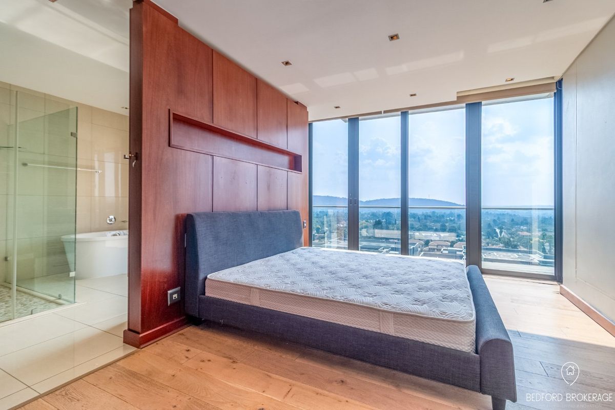 Modern bedroom featuring floor-to-ceiling windows with panoramic views, hardwood flooring, and an integrated open-plan bathroom with a glass shower and freestanding tub.