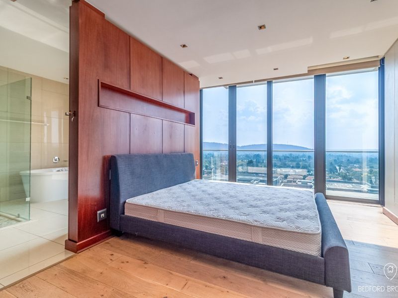 Modern bedroom featuring floor-to-ceiling windows with panoramic views, hardwood flooring, and an integrated open-plan bathroom with a glass shower and freestanding tub.