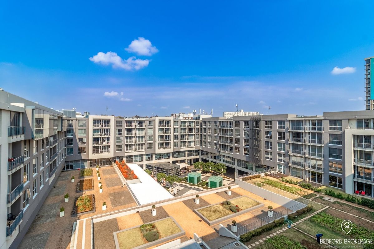 An elevated aerial view of the apartment complex courtyard, featuring landscaped garden beds, paved pathways, and surrounding residential building facades.