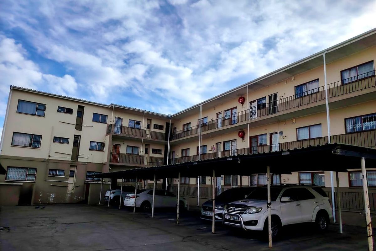 Exterior view of an apartment complex featuring multi-level balconies, metal railings, and a covered parking area with concrete paving.