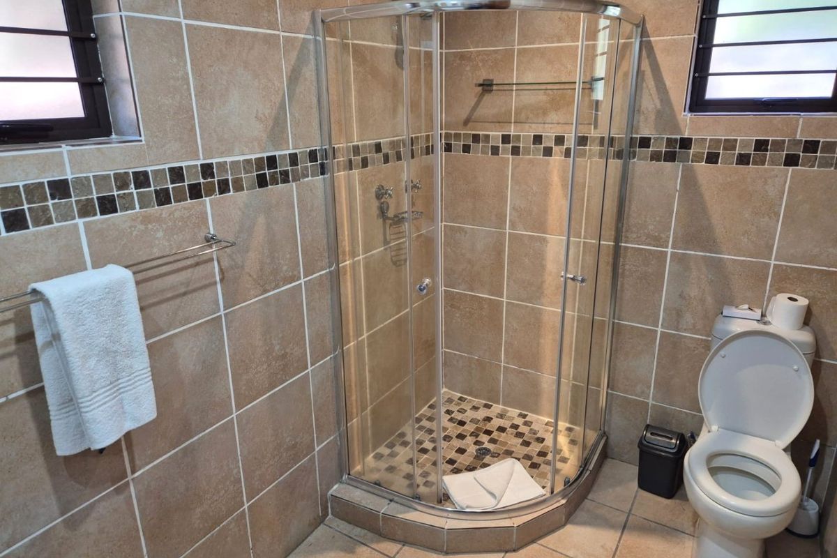 A functional bathroom featuring a curved glass shower enclosure, ceramic wall and floor tiling, and a standard toilet with louvered windows.