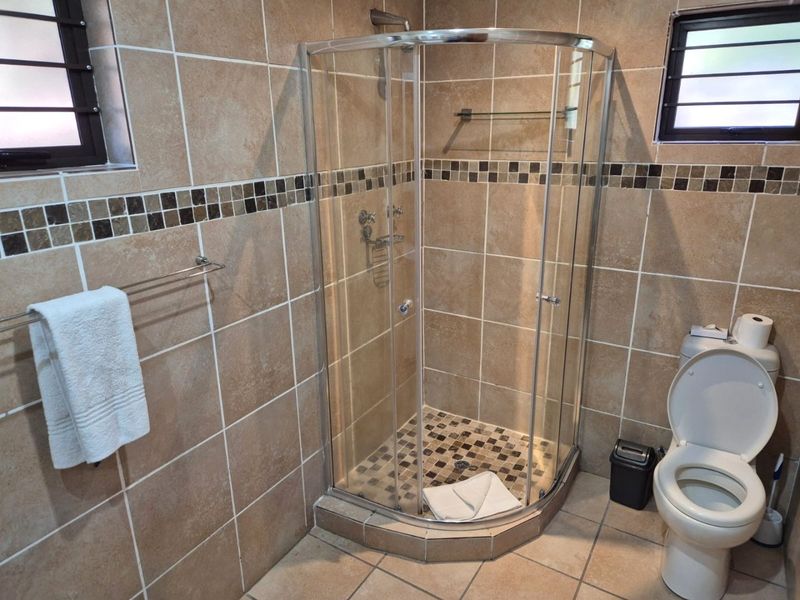 A functional bathroom featuring a curved glass shower enclosure, ceramic wall and floor tiling, and a standard toilet with louvered windows.