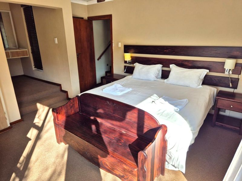 A spacious bedroom featuring a built-in wooden headboard, wall-mounted lighting, and carpeted flooring, with an adjacent elevated area and internal staircase access.