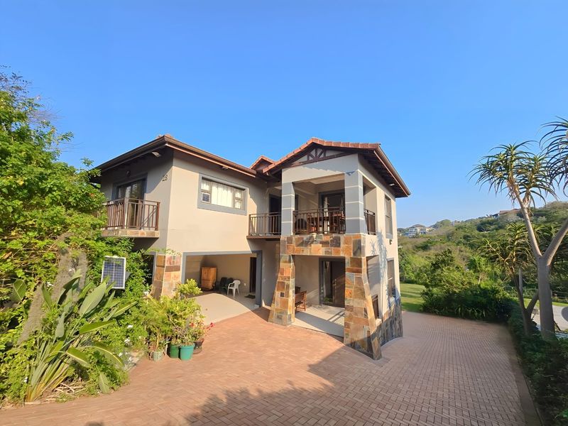 A multi-level residential property featuring stone-clad pillars, a paved driveway, and balconies overlooking a landscaped garden area.
