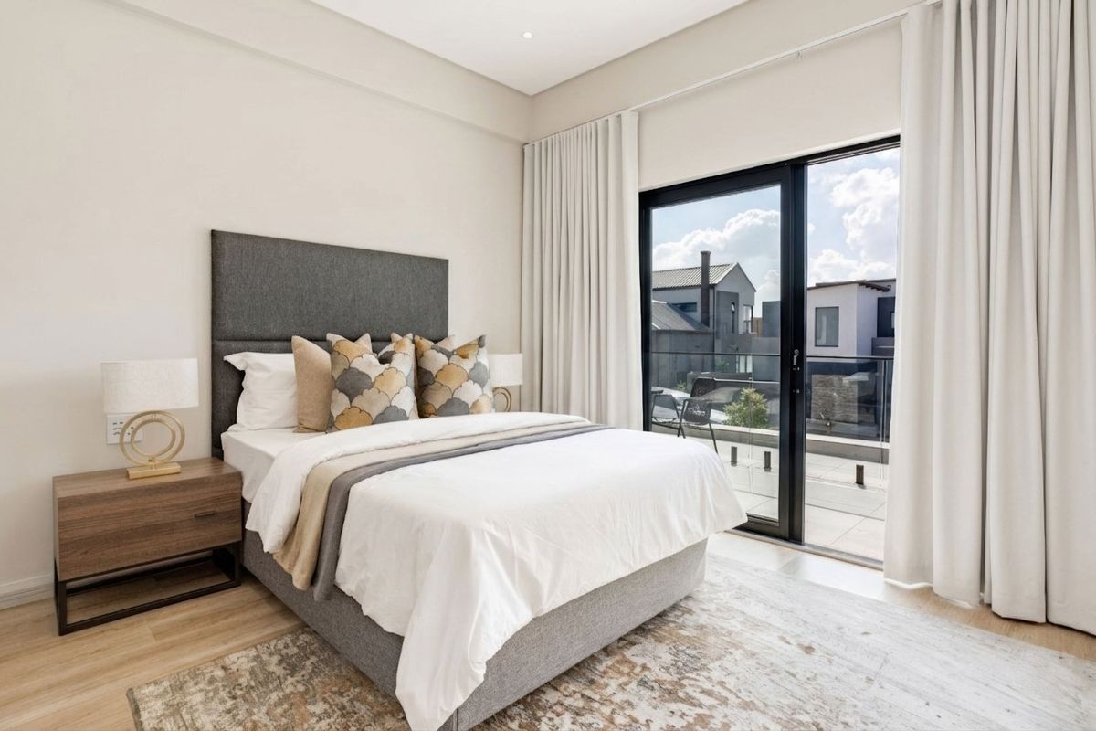 A modern, well-lit bedroom featuring neutral finishes, laminate flooring, and a large sliding glass door providing direct access to a private balcony.
