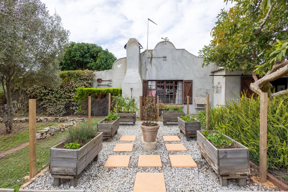 Rear view of a farm-style cottage featuring a gabled roofline, chimney, and a landscaped garden area with gravel paths and raised timber garden beds.