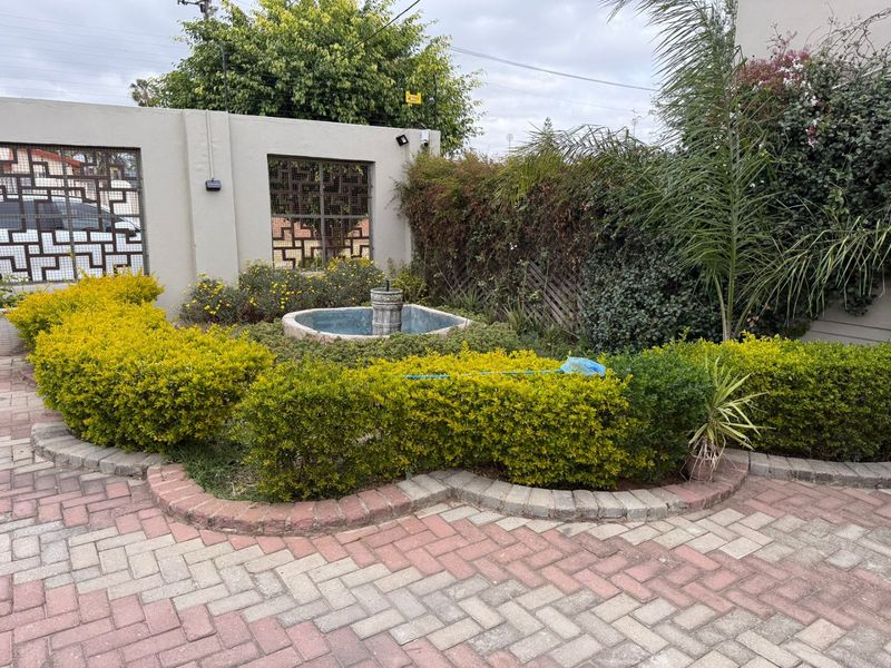A front garden area featuring a paved driveway, boundary wall with decorative metal screens, and a central water fountain surrounded by established shrubbery.