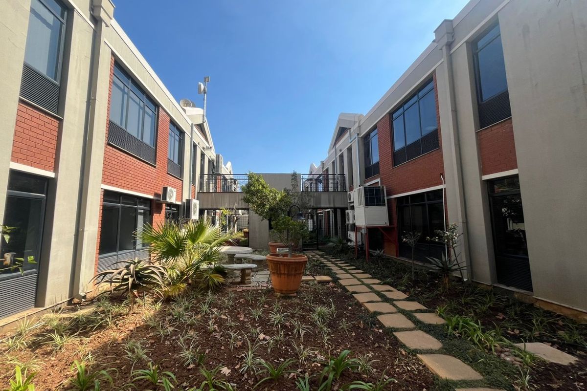 A landscaped courtyard area between two commercial building wings, featuring a paved walkway, established garden beds, and an elevated pedestrian bridge connecting the structures.
