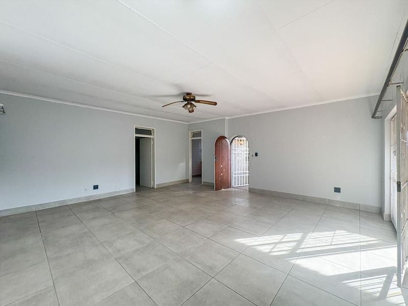 A spacious, empty living area featuring neutral tiled flooring, a ceiling fan, and an arched entrance with a security gate.
