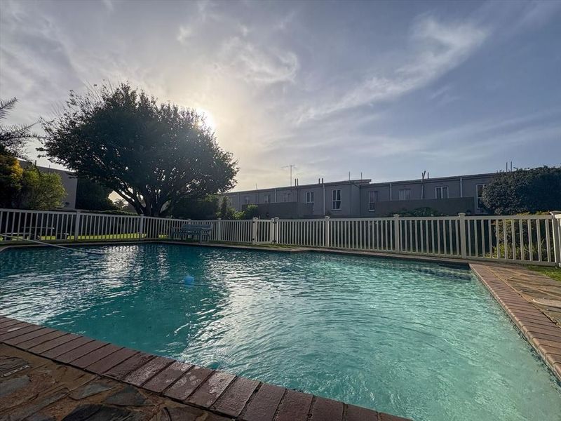 A well-maintained swimming pool area featuring brick coping and a white picket fence, set within a communal townhouse complex garden.