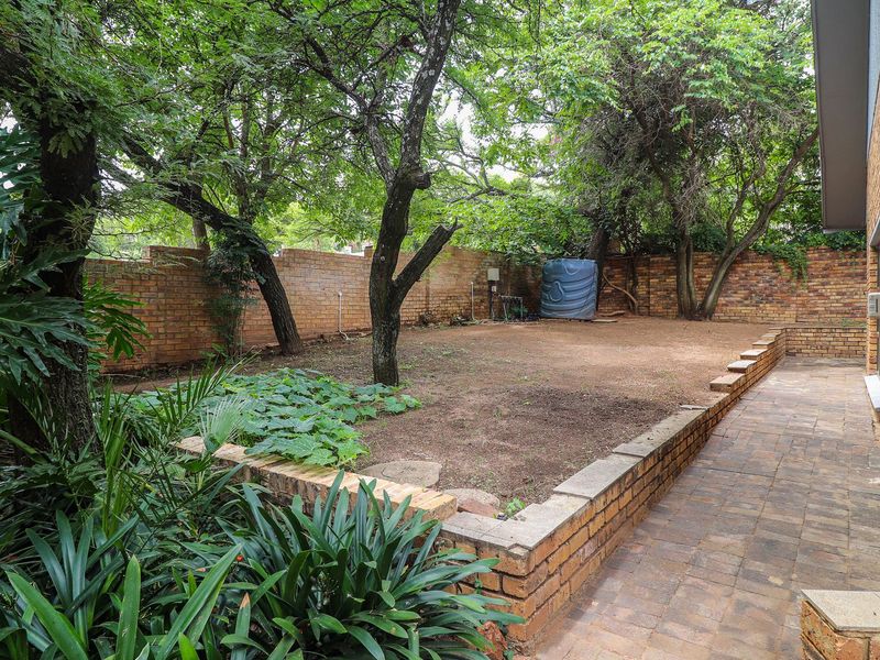 A private rear garden featuring a paved patio area, brick retaining walls, mature trees, and a perimeter brick boundary wall.
