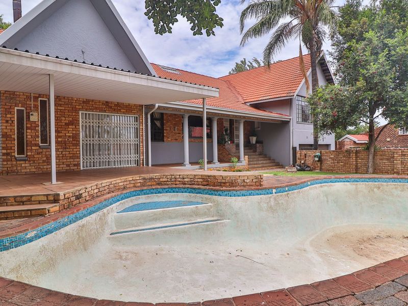 Rear exterior view featuring a large swimming pool, covered patio area, and brick boundary wall with traditional terracotta roof tiles.