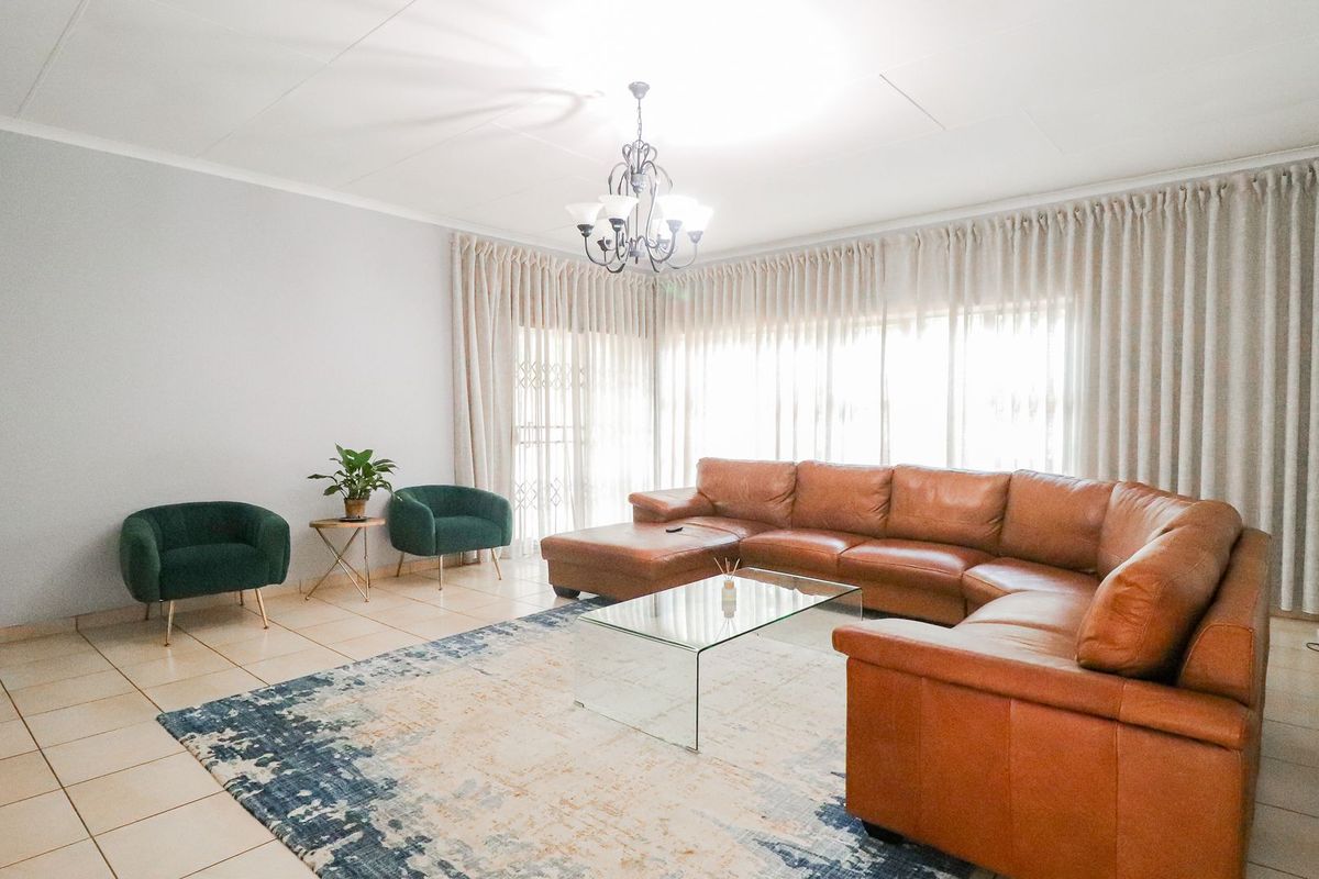 A spacious living room featuring neutral tiled flooring, a central chandelier, and large windows dressed with floor-to-ceiling curtains.