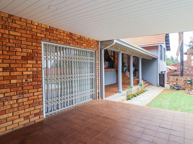 A spacious covered patio area featuring tiled flooring, brick wall finishes, and a security gate, overlooking a well-maintained garden and exterior walkway.