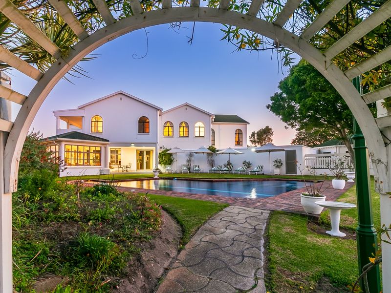 A beautifully landscaped backyard featuring a swimming pool, brick-paved surround, and a charming arched pergola framing the view of the main building structure.