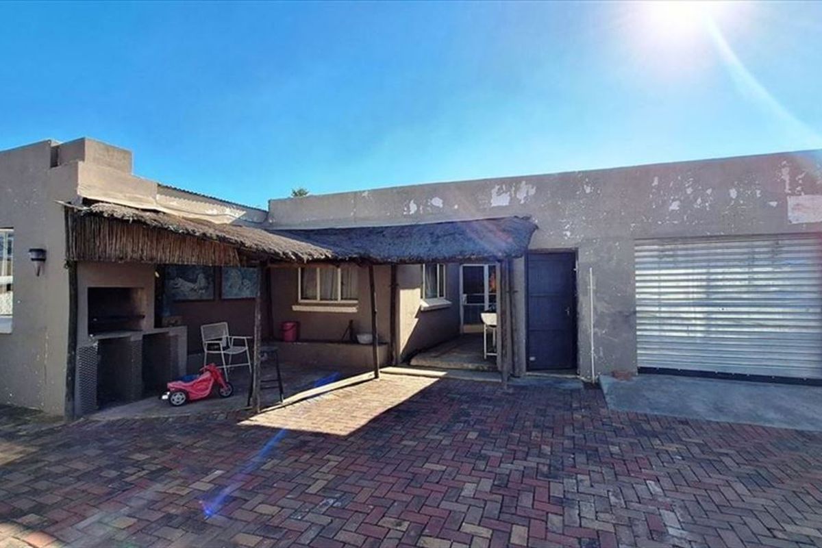 Rear exterior view featuring a paved courtyard, a built-in braai area with a thatched roof, and a garage with a roller shutter door.