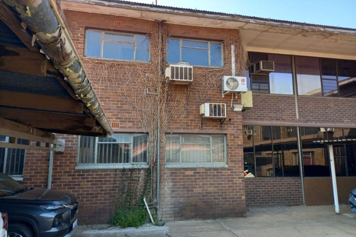 Exterior view of a commercial brick building featuring multiple wall-mounted air conditioning units, metal-framed windows, and a covered parking area.
