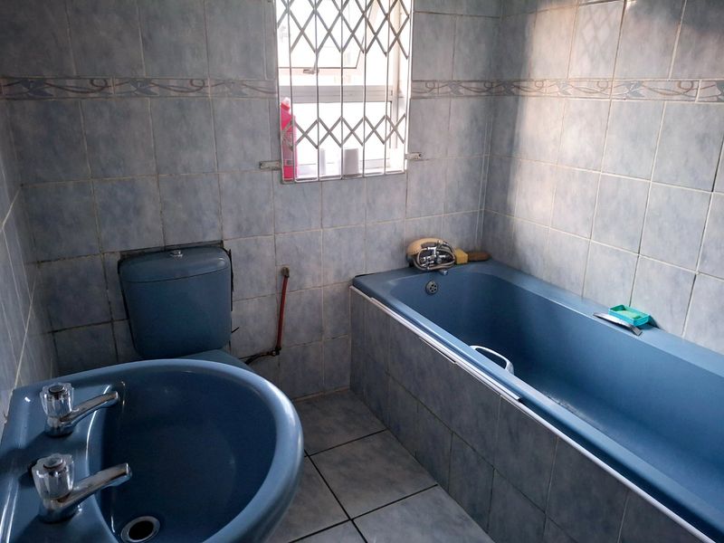 A dated bathroom featuring blue sanitaryware, tiled walls and flooring, and a window fitted with a security gate.